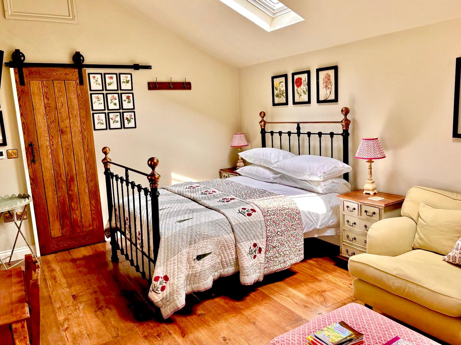 The bedroom: a brass and iron bed with a floral quilt beneath a skylight, a pale yellow armchair, and the sliding oak door to the shower room