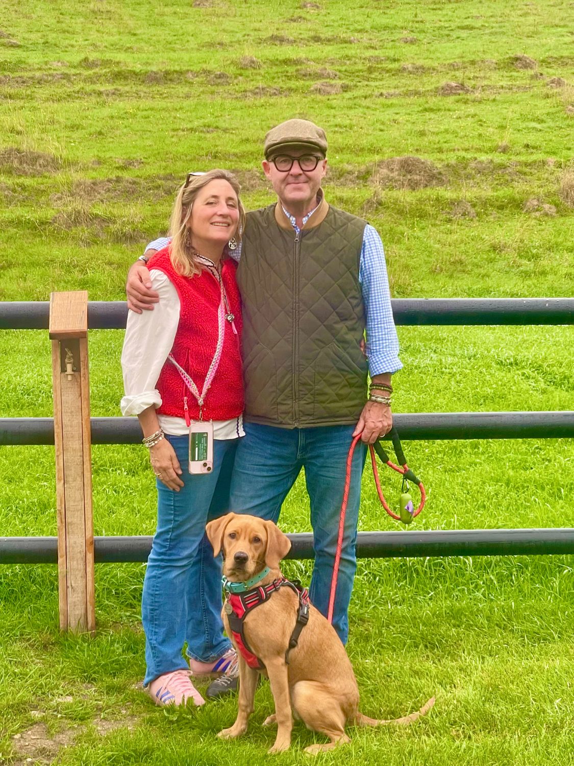 Your hosts Vics and Adam standing by a paddock fence with Suki, their fox‑red Labrador