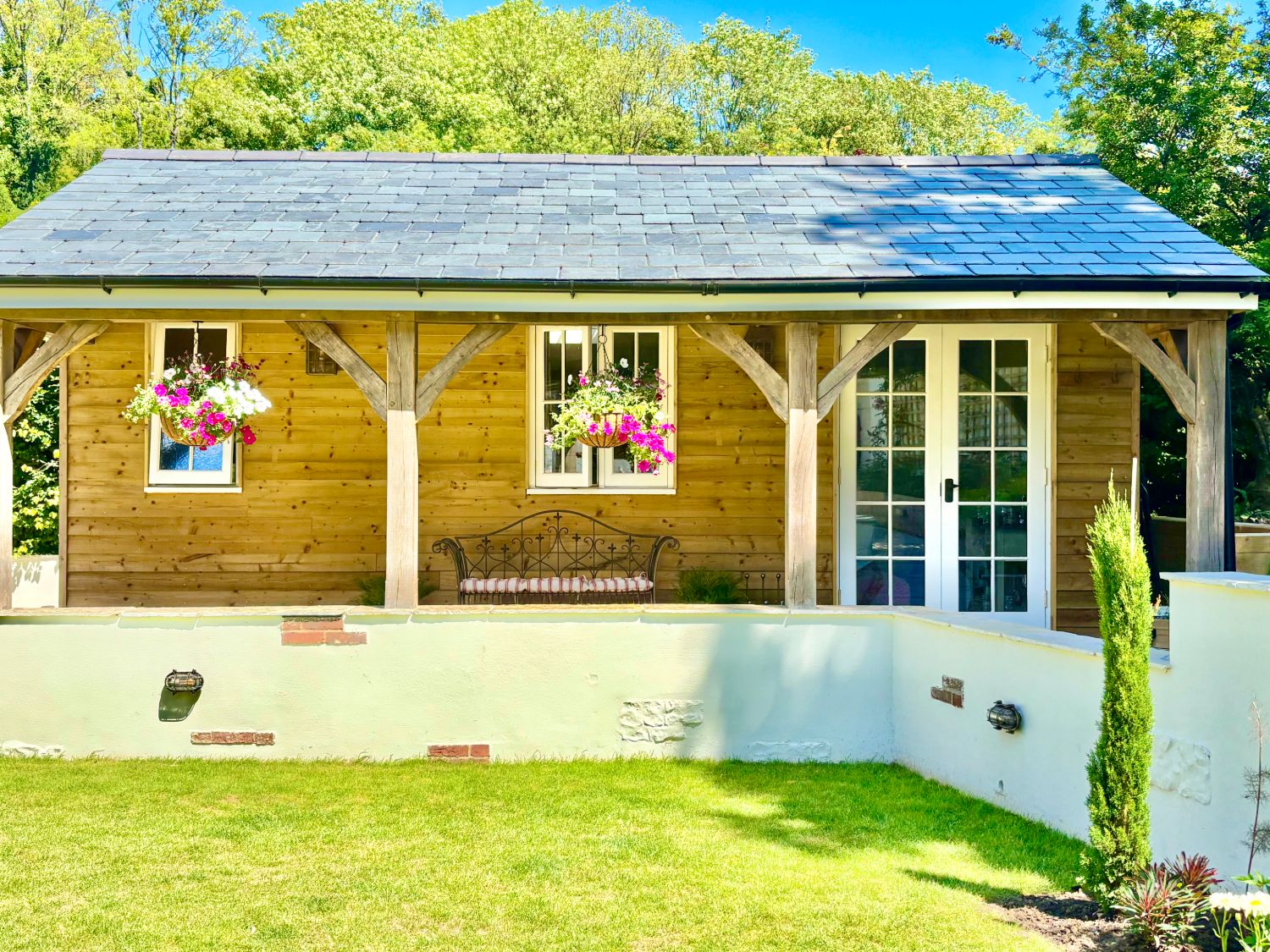 Garden Lodge seen from the lawn: larch cladding, an oak‑framed veranda with hanging baskets, a garden bench and French doors